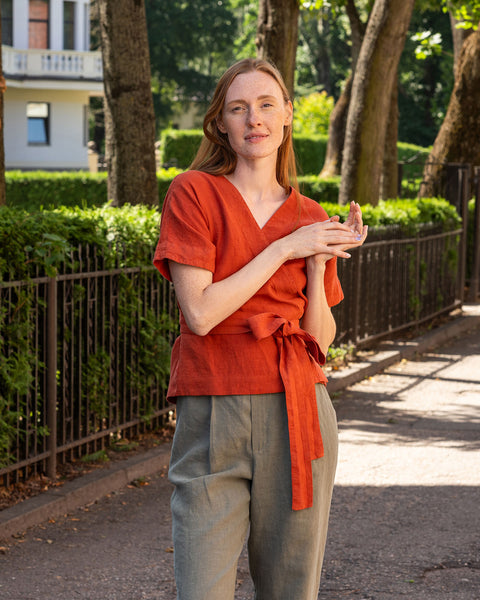 A person with light skin and dark hair stands in profile, looking down towards their hands which are crossed at their midsection. They are wearing a linen wrap top with short, loose sleeves and stone green wide-leg pants. In the background, there is a dark metal fence and lush green foliage.