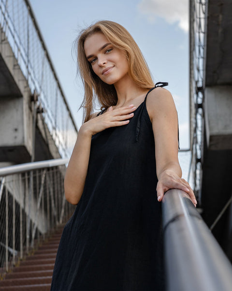 A light-skinned person with long blonde hair is standing on a set of outdoor stairs, looking towards the camera with a gentle smile. They are wearing a pure black sleeveless linen dress with thin straps, and one hand is resting on their chest while the other holds onto a metal railing. The background shows a cloudy sky and architectural elements of a walkway or bridge.