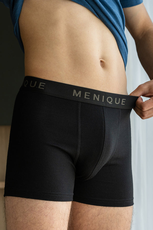 a man wearing short boxer briefs made from merino wool fabric, in color black. He is pulling the logo waistband, to show the stretch of the garment.