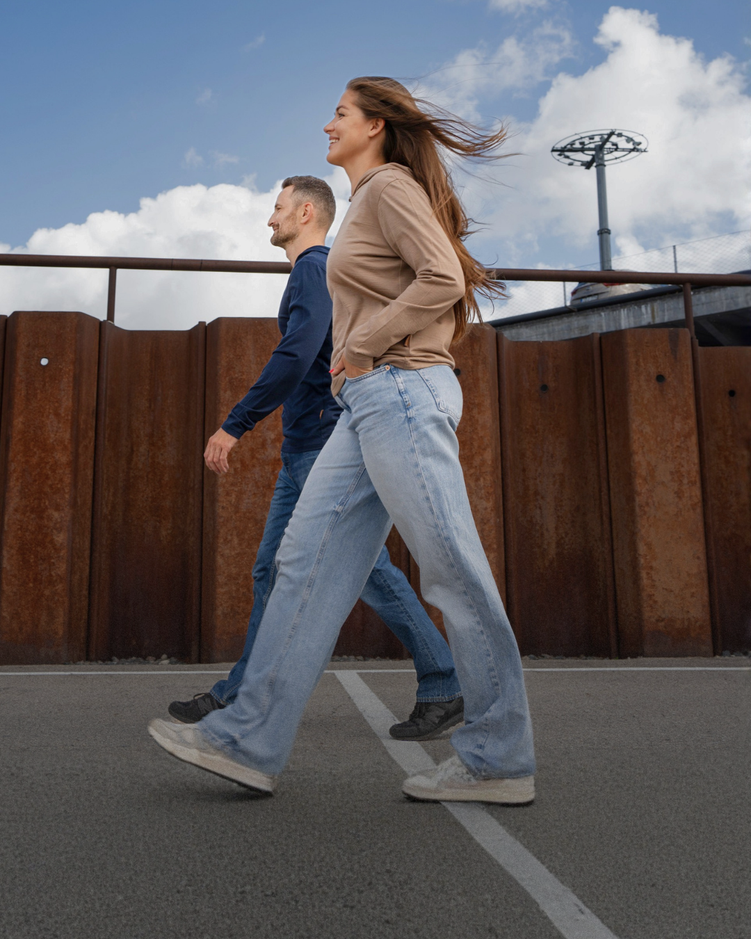 A woman in a beige cardigan and light blue wide-leg jeans walks alongside a man in a navy blue shirt and jeans on an outdoor paved walkway with a rusted metal fence and blue sky in the background.