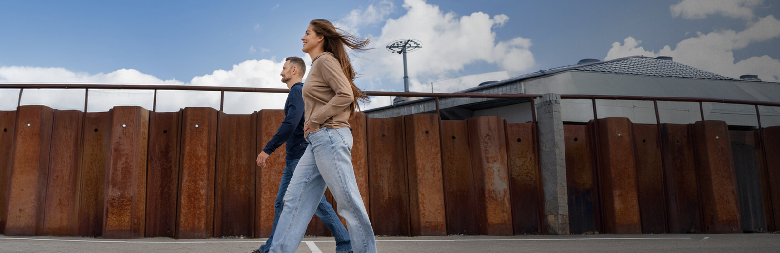 A woman in a beige jacket and light blue jeans walks beside a man in a dark blue shirt and jeans along an outdoor walkway with a tall rusted metal fence, blue sky, and buildings in the background.