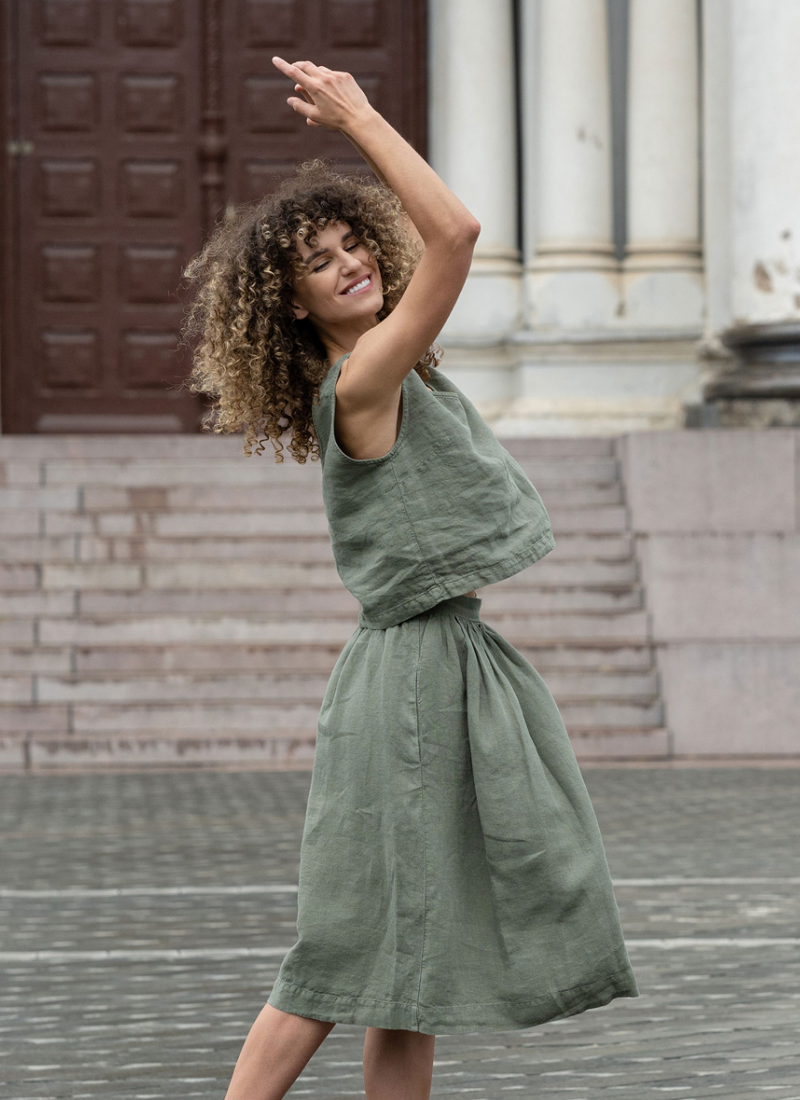 A smiling woman with curly hair is captured mid-spin in a stone-paved public square, her eyes closed and arms raised gracefully above her head. She wears a matching olive green linen set consisting of a loose sleeveless top and a flared midi skirt, with stone steps and classical building columns in the background.