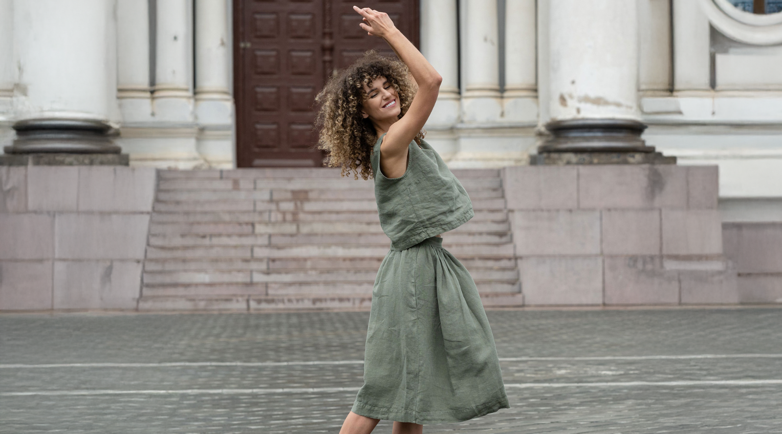 A smiling woman with curly hair is captured mid-spin in a stone-paved public square, her eyes closed and arms raised gracefully above her head. She wears a matching olive green linen set consisting of a loose sleeveless top and a flared midi skirt, with stone steps and classical building columns in the background.