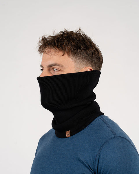 A person with short brown hair is shown in profile, wearing a black knit neck gaiter pulled up to cover their nose and mouth. They are wearing a blue long-sleeved shirt. The background is a plain light gray. A small brown tag is visible on the neck gaiter.
