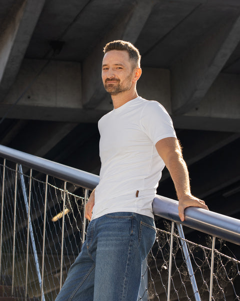 A man leans gently against a metal wire railing on an outdoor staircase, positioned underneath a large concrete bridge structure. He is wearing a plain white short-sleeved t-shirt and blue jeans, looking directly at the camera with a subtle smile.