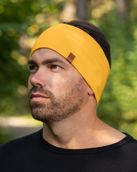A man with short, dark hair and a beard is shown from the chest up, wearing a spicy yellow merino wool headband with a small brown tag. He is wearing a black shirt and looking off to his left with a neutral expression. The background is softly blurred, showing green foliage and tree trunks, suggesting an outdoor setting.