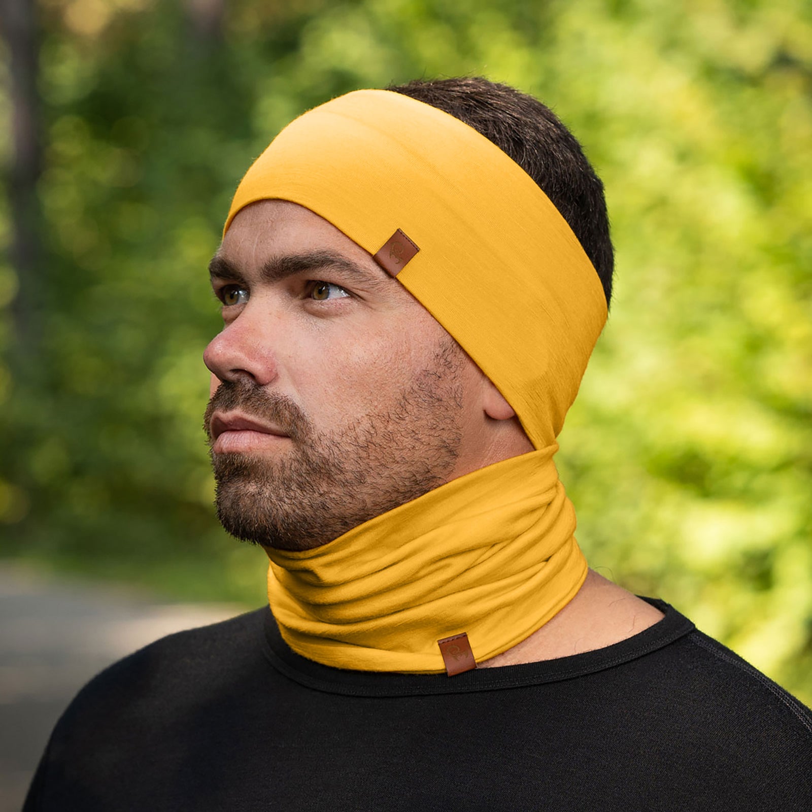 A person with short, dark hair is shown wearing a spicy yellow headband and a matching neck gaiter that is pulled up to cover their mouth and nose, with only their eyes visible. They are wearing a black long-sleeved shirt, and the blurred background suggests an outdoor setting with green foliage.