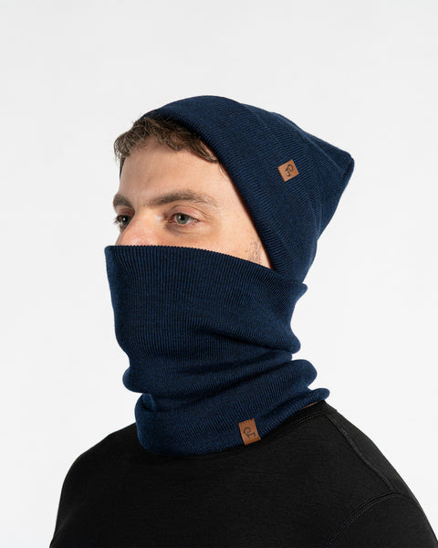 A male model, viewed in profile, wears a blue beanie and a matching neck gaiter that covers his nose and mouth. Both the beanie and gaiter feature a small, brown leather patch with a Menique logo. He has a short beard and dark hair. The background is solid white.