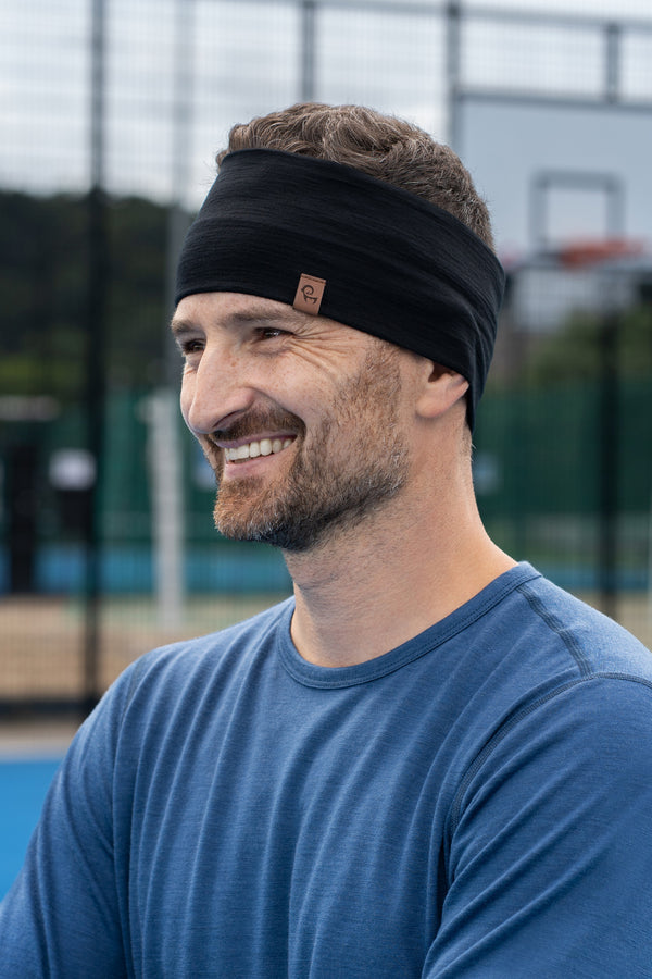 A close-up shot of a smiling man looking to the side while standing on a bright blue outdoor sports court. He is wearing a denim-blue long-sleeved shirt and a wide black headband. The background features a blurred chainlink fence and a basketball backboard.