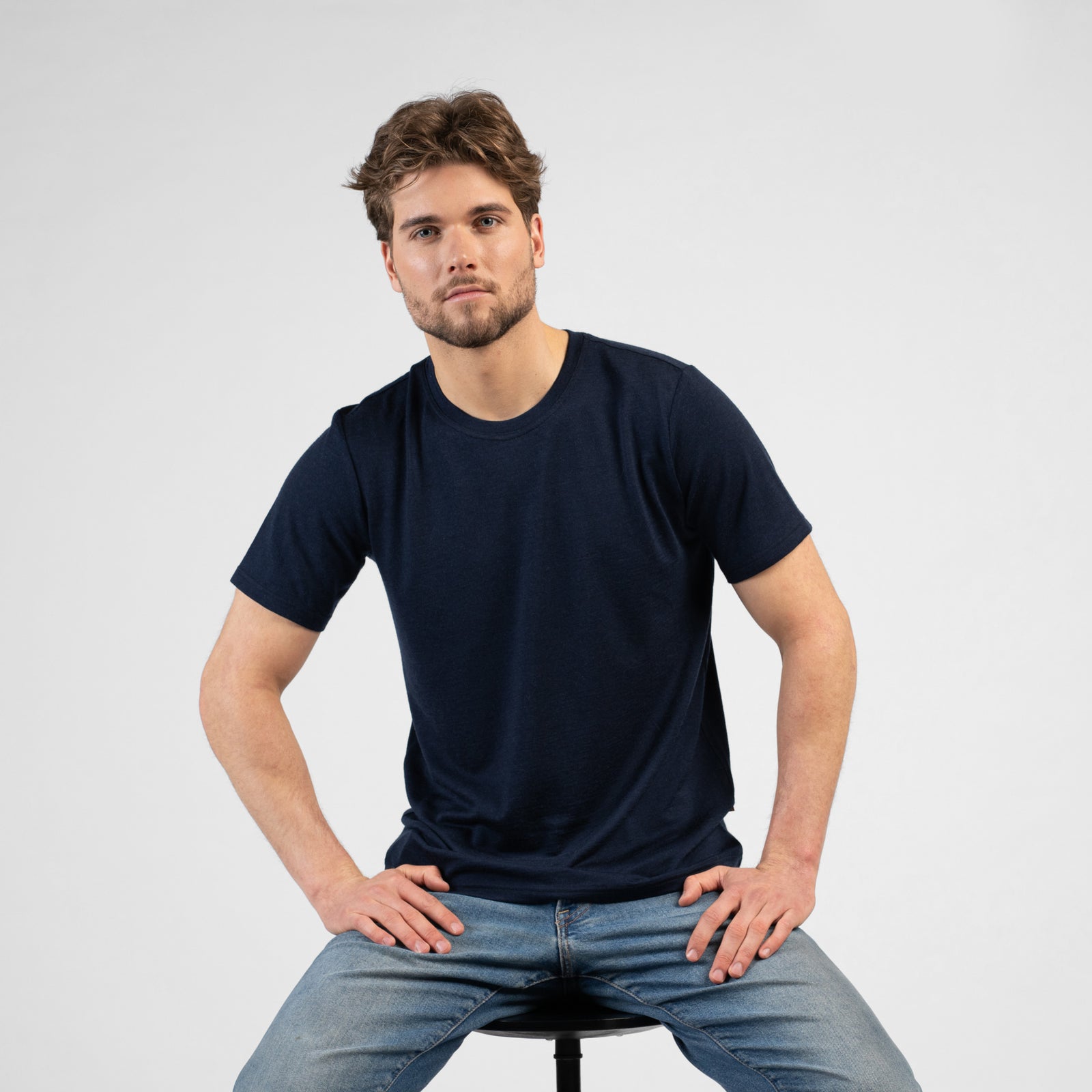 A smiling man standing outdoors in front of rusty metal pillars. He is wearing a navy blue short-sleeve crew neck t-shirt and beige shorts, with his right hand casually resting in his pocket as he looks off into the distance.