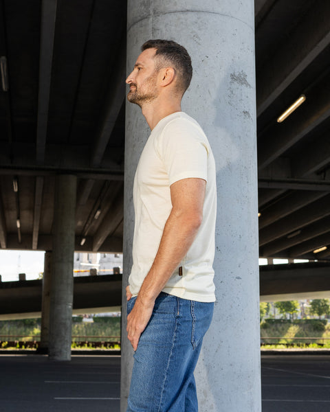 A side profile view of a man standing next to a large concrete pillar underneath an overpass. He is wearing a short-sleeved, natural off-white crew neck t-shirt and blue jeans, looking straight ahead.