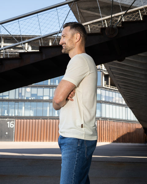 A man stands with his arms crossed, smiling and looking off to the left. He is wearing a short-sleeved, natural off-white crew neck t-shirt and blue jeans, positioned in an outdoor space with modern architecture and a rusted steel structure behind him.