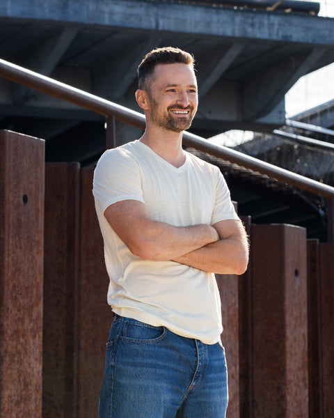 A smiling man stands outdoors with his arms crossed over his chest, looking off into the distance. He is wearing a short-sleeved, natural off-white v-neck t-shirt and blue jeans, with a backdrop of rusted steel structures and a concrete bridge.