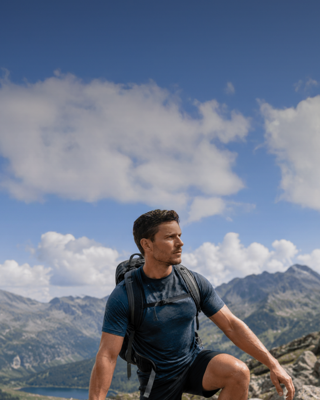 A man wearing a dark blue short-sleeve t-shirt, black shorts, and a black backpack stepping up a rocky mountain trail, with a distant blue lake and mountain peaks in the background.