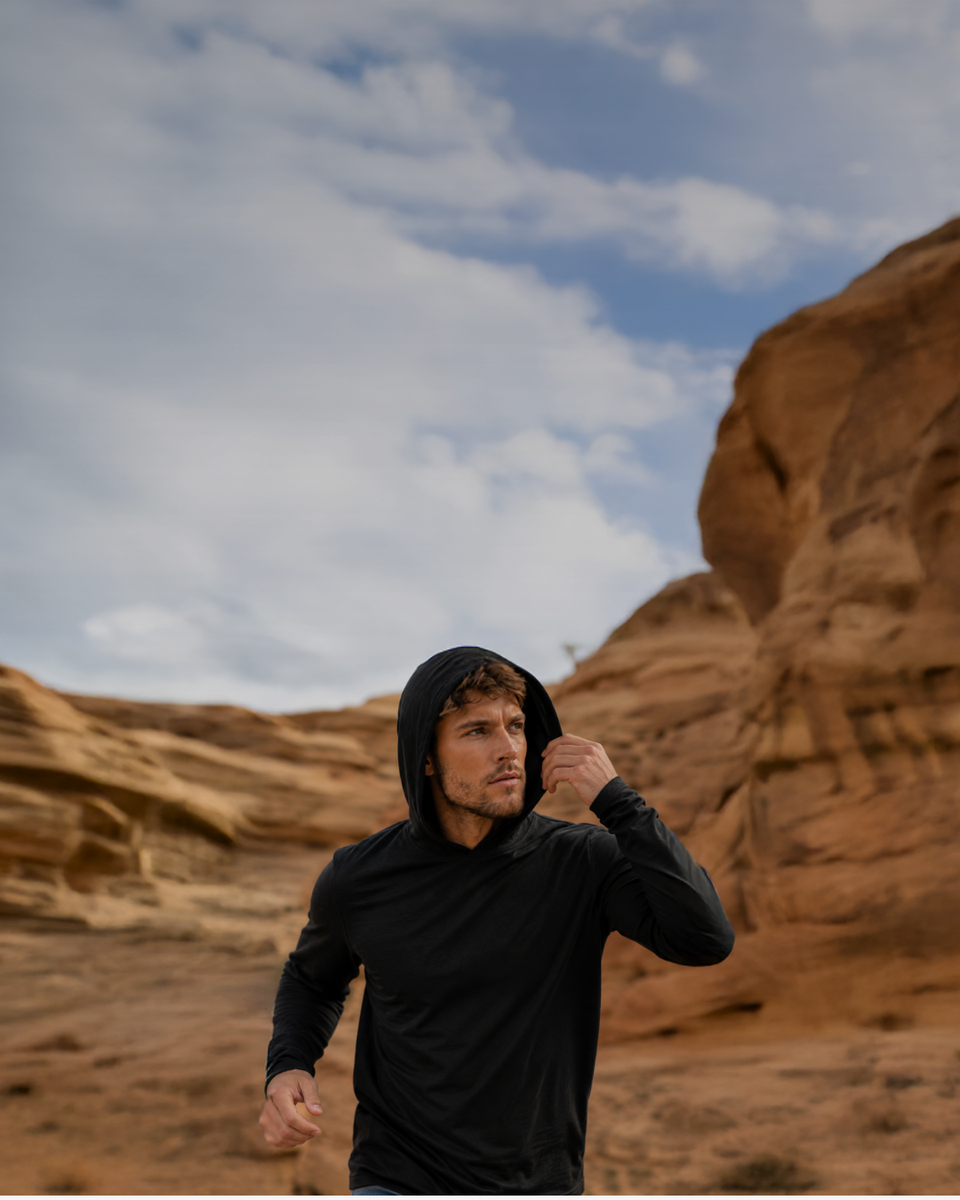 A man wearing a black long-sleeve sun hoodie adjusting his hood while standing in a rugged, wind-swept rocky desert landscape.