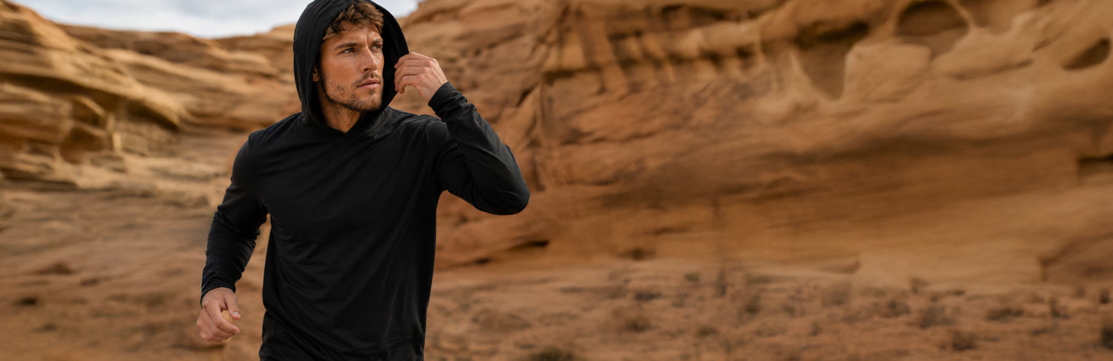 A man wearing a black long-sleeve sun hoodie adjusting his hood while standing in a rugged, wind-swept rocky desert landscape.