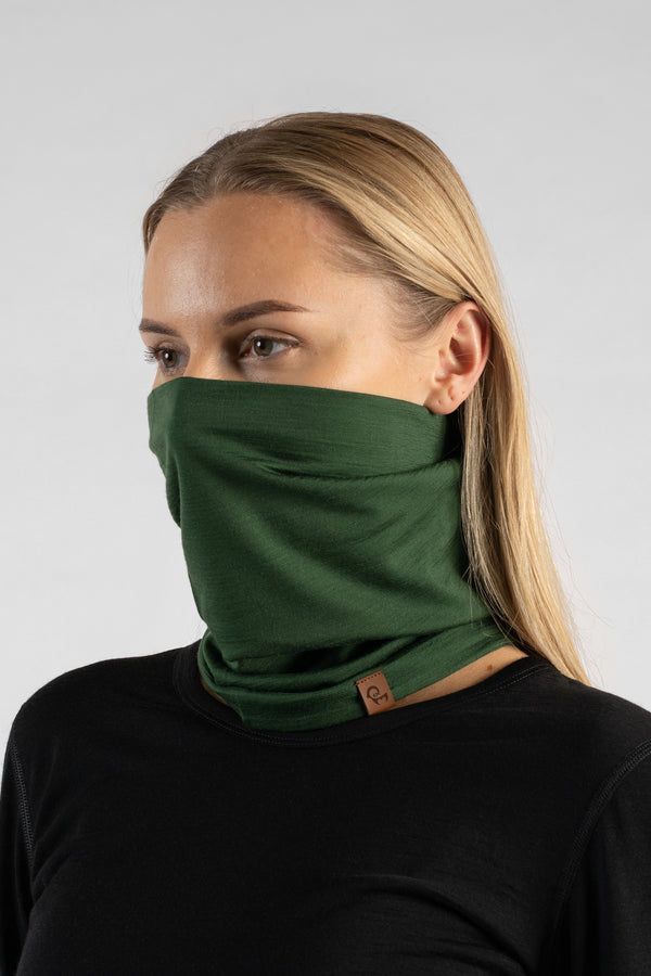 A studio portrait of a blonde woman looking off to the left against a plain light grey background. She is wearing a black long-sleeved shirt and a dark green neck gaiter pulled up to cover her nose and mouth, featuring a small brown logo tag at the bottom.