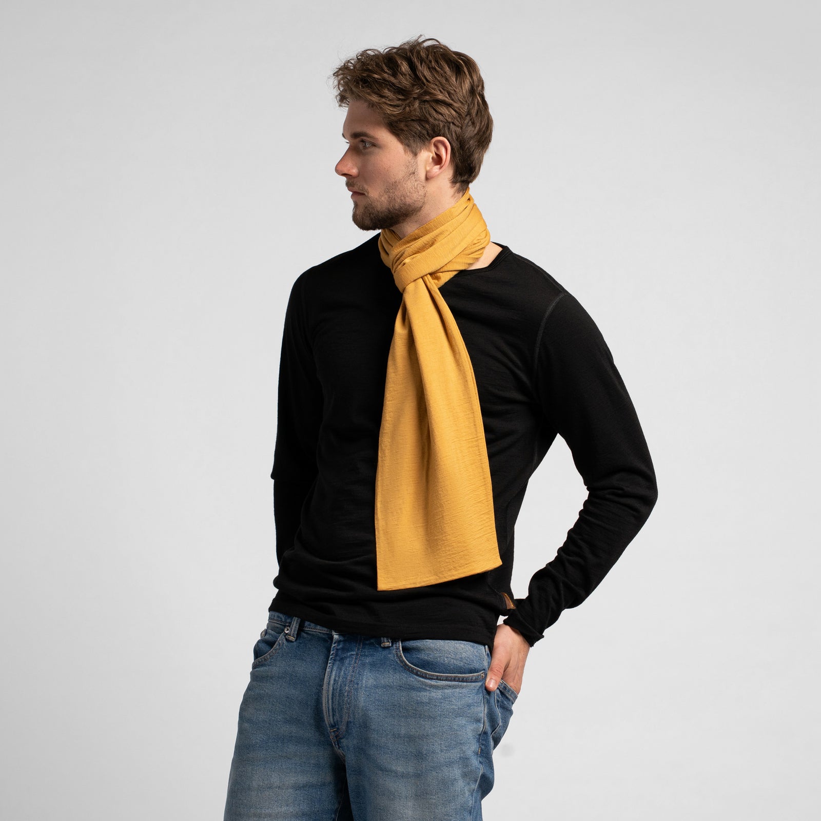 A studio portrait of a man looking off to his left, with his hands in his blue jeans pockets. He is wearing a fitted black long-sleeved shirt and a mustard yellow scarf wrapped once around his neck. The scarf's ends drape down his chest, and a small, square brown leather logo patch is visible on the bottom edge of one side. The background is a solid light grey.