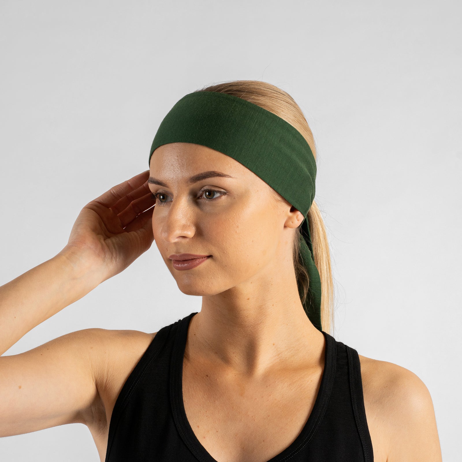 A studio portrait of a woman looking downwards and to the right as she ties a solid black headband behind her head. Her blonde hair is pulled back into a ponytail, and she is wearing a black tank top. The background is a solid light grey.