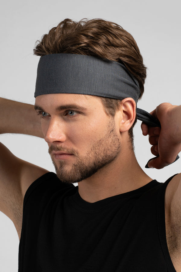 A man with wavy brown hair looks to the left while tying a grey headband behind his head. He is wearing a black tank top against a plain light grey background.