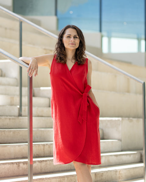 A woman with brown hair, wearing a bright red, sleeveless wrap dress, poses on indoor concrete stairs with metal railings.
