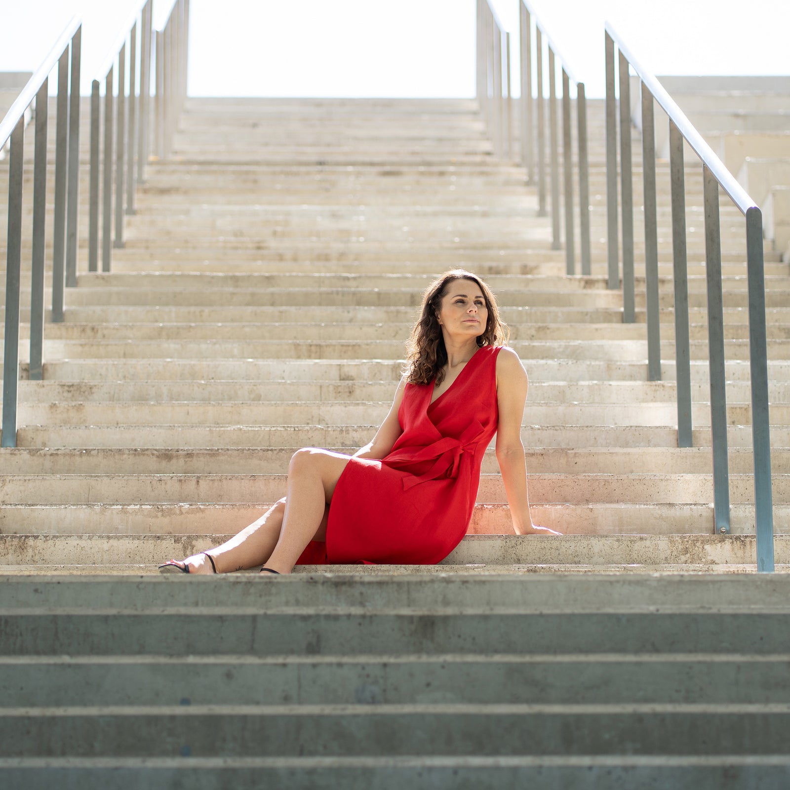 A woman with brown hair, wearing a bright red, sleeveless wrap dress, poses on indoor concrete stairs with metal railings.
