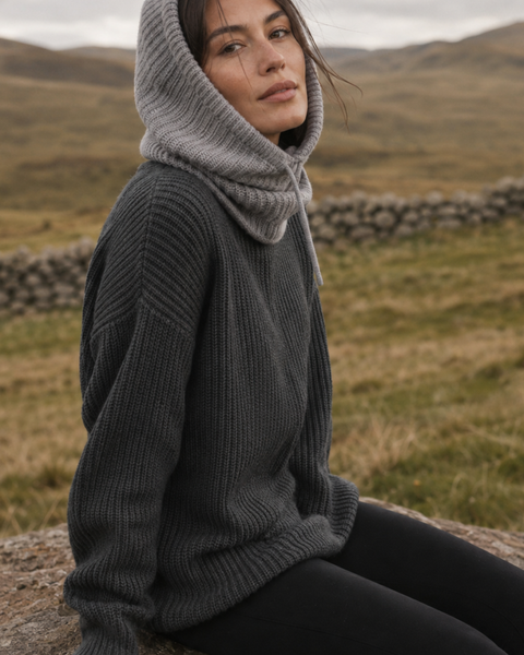 A medium shot of the woman sitting on a large rock in the same hilly landscape. She is wearing the light grey knit balaclava, dark grey sweater, and black leggings. Her body is angled slightly to the side while she looks back toward the camera.