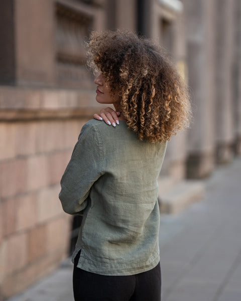 Woman with curly hair wearing a green linen blouse walking away from the camera on a city street.