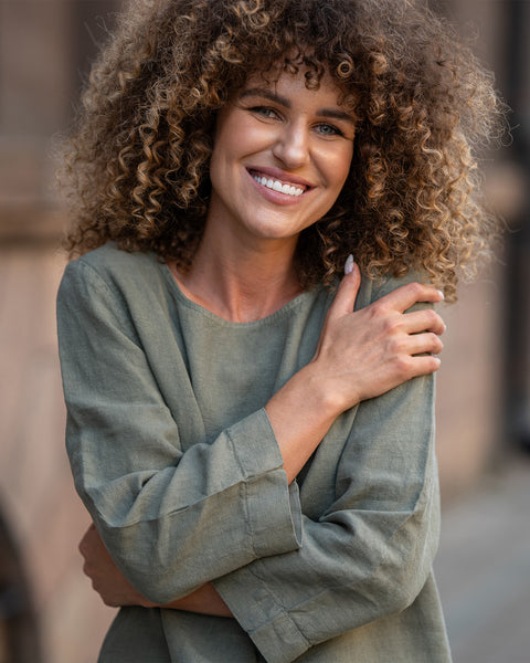 Woman with curly hair wearing a green linen blouse, smiling outdoors.