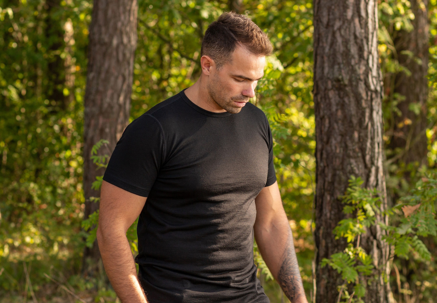 A man wearing a fitted black merino wool T-shirt from menique walks outdoors in a forest setting, with trees and sunlight in the background.
