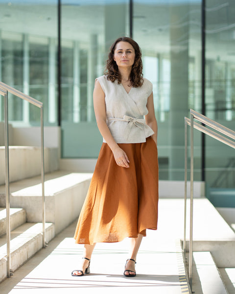 A woman with brown hair, wearing a light beige wrap top and a long rust-brown skirt, poses indoors in front of large windows.
