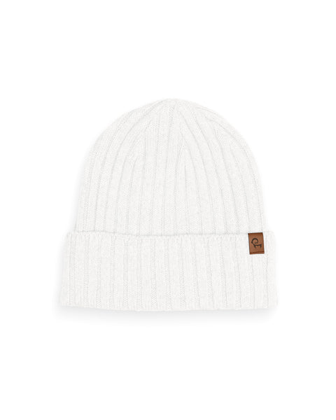 A white ribbed knit beanie is displayed against a white background. The beanie has a folded brim and a small brown square tag is attached to the brim on the right side. The knit has vertical ribbing.