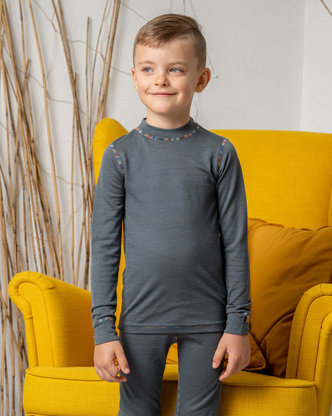 A boy standing in front of a yellow armhair, smiling and looking to the side. He is wearing the menique 160gsm merino long sleeve top in perfect gray color, with matching pants.