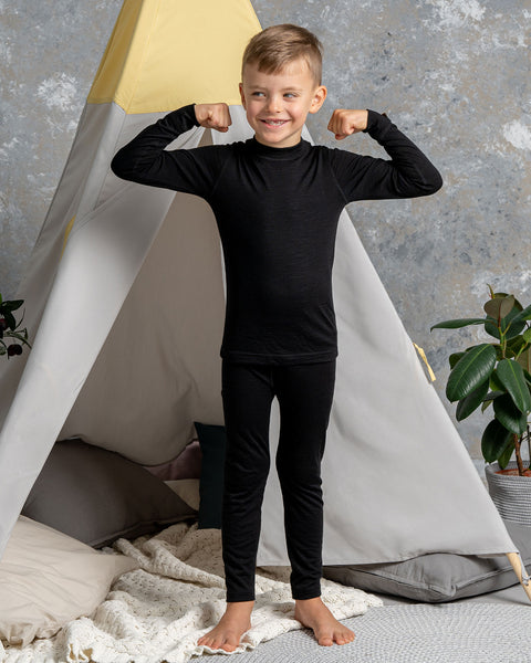 a boy wearing the menique Kids&#39; 160 Thermal Long Sleeve Crew in Black color. He has his arms raised and bent, acting strong. He is barefoot on a carpet.