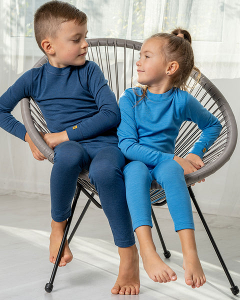 Two children, a boy and a girl, with the girl wearing the menique Kids&#39; 160 Merino Pants in Light Blue color, and the boy the same, yet in denim color. Both of them are sitting in a lounge chair. 