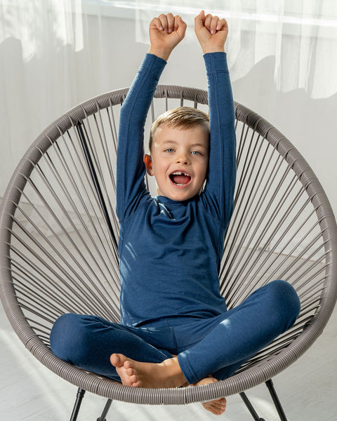 A boy sitting in a lounge chair, wearing the menique Kids&#39; Merino 160 Long Sleeve Set in Denim color. 