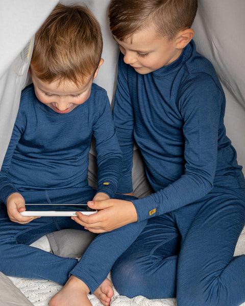 Two boys wearing the menique merino 160gsm long sleeve set in denim color. They are playing a mobile game with a phone, in a children&#39;s indoor play-tent.