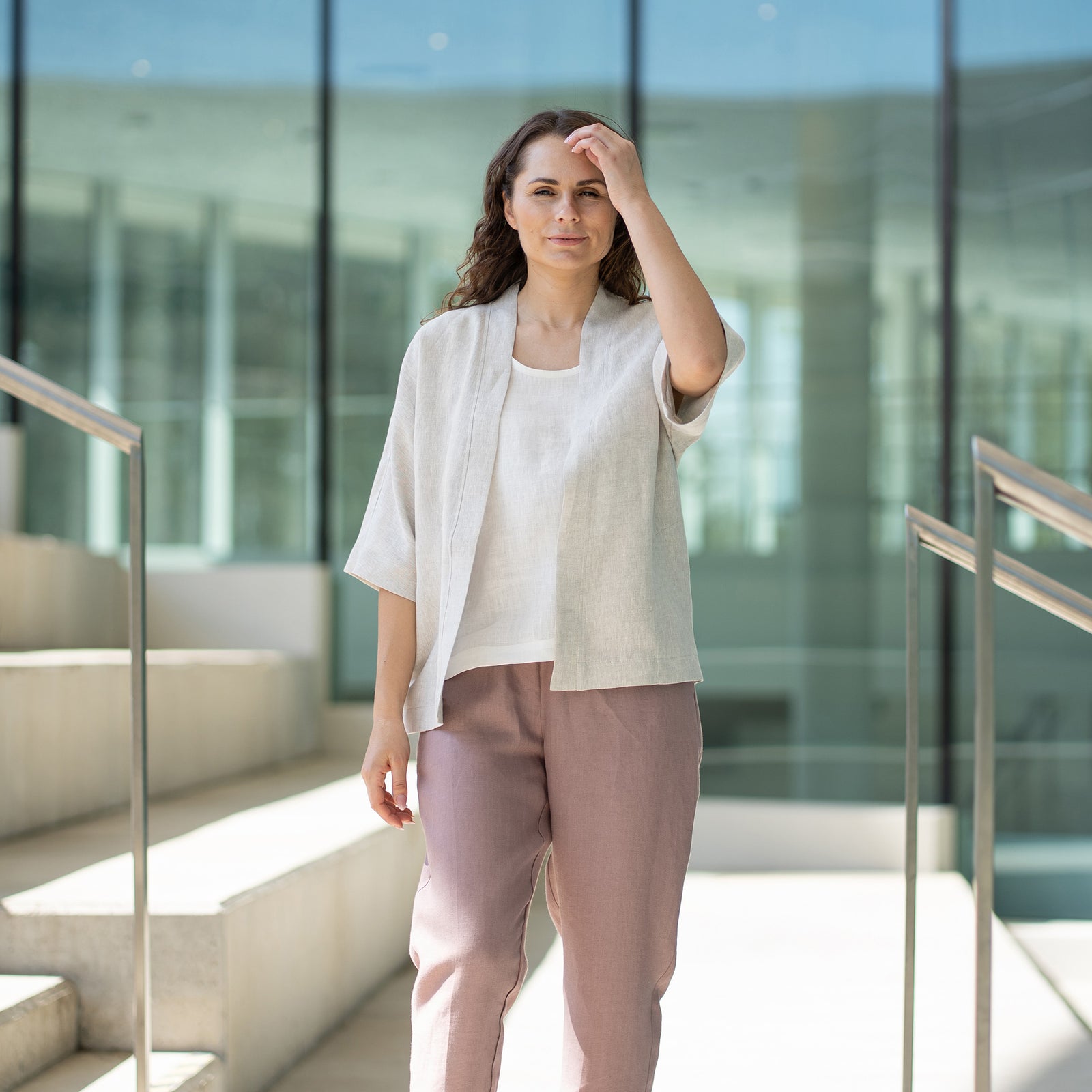 A woman with brown hair, wearing a light beige kimono-style jacket over mauve trousers, stands on indoor concrete stairs.
