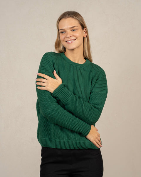 A woman, hugging herself, looking to the side and smiling. She is wearing the menique combed cotton sweater in green color.