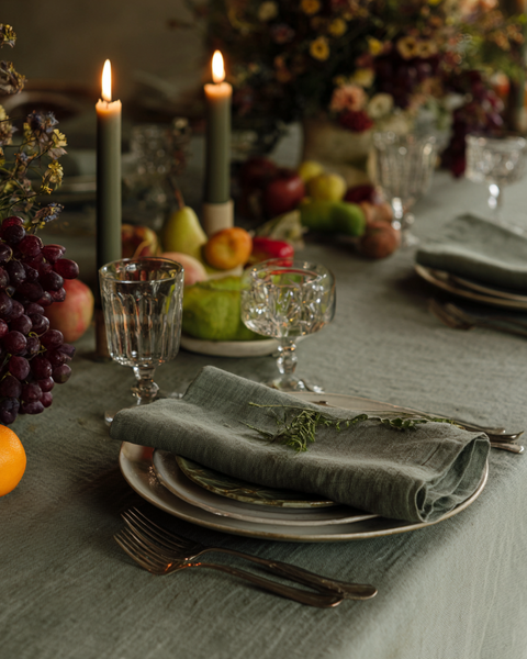 Rustic fall table setting with sage green linen tablecloth, matching linen napkins, candles, and harvest fruits for an elegant holiday tablescape.