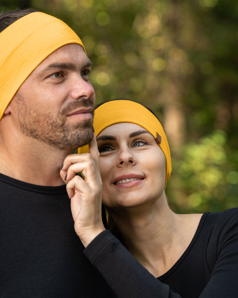 A man and a woman are closely positioned outdoors, both wearing spicy yellow menique merino wool headbands and black long-sleeved shirts. The man, on the left, has short brown hair and a beard and is looking upwards and to his left. The woman, on the right, has light brown hair and is looking upwards with a smile, her hand gently touching the man&#39;s face. The background is softly blurred with green and brown tones, suggesting a natural, possibly wooded, environment.
