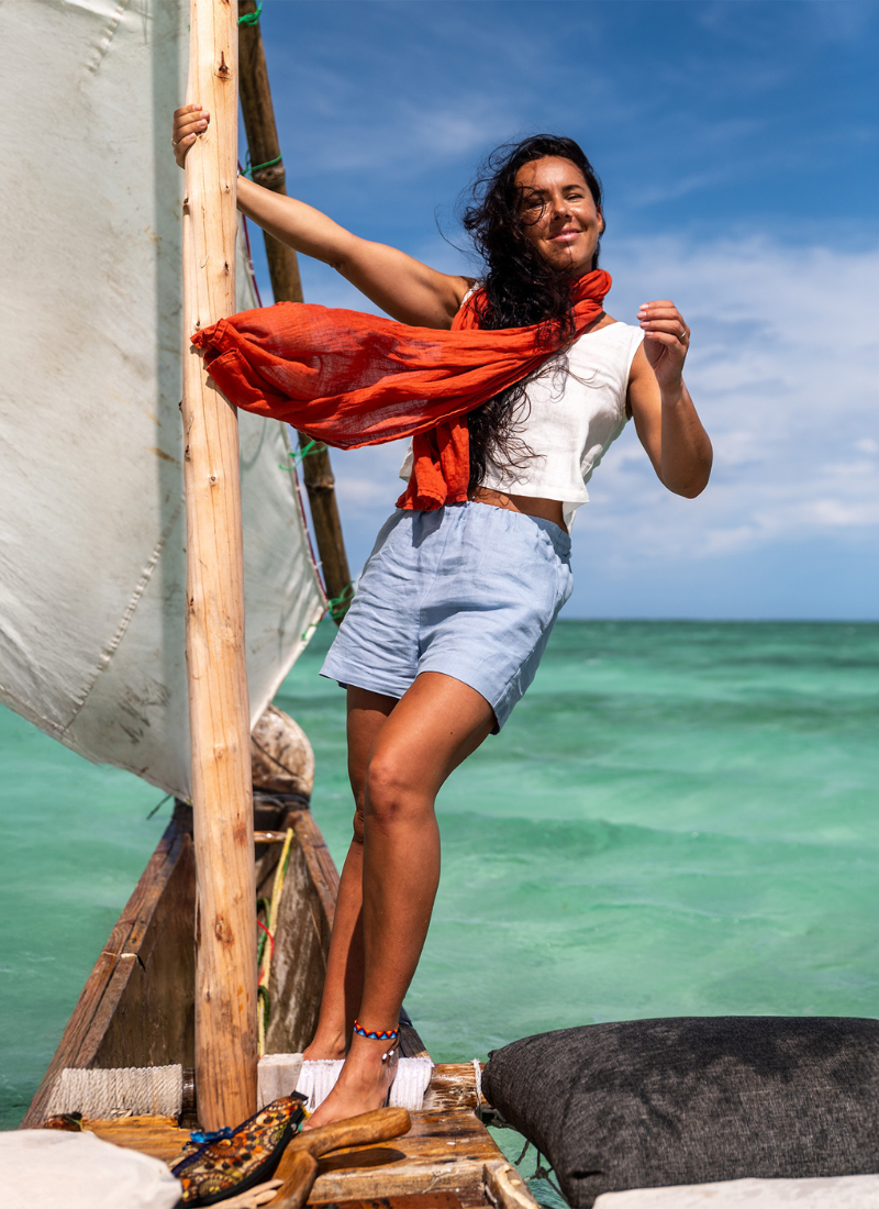 Woman wearing a white sleeveless top and light blue linen shorts standing on a wooden boat, with a flowing red scarf and turquoise sea in the background, capturing a relaxed summer vacation mood by the ocean.