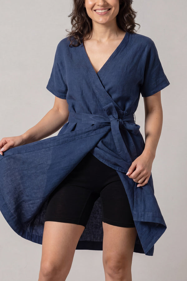 A smiling woman wearing a short-sleeved, navy blue linen wrap dress holds the front of her skirt open to reveal fitted, mid-thigh black slip shorts worn underneath.
