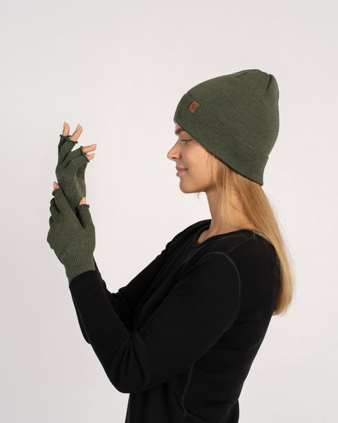 Side view of a woman wearing a dark-green knit beanie and matching fingerless gloves, inspecting her raised hands.