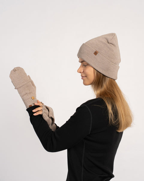 Side-profile of a woman in a creamy-beige beanie and mittens, hands raised as she adjusts the glove flaps.