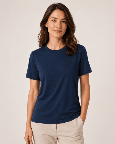 A brunette woman with shoulder-length wavy hair modeling a navy blue, short-sleeve crew neck t-shirt. She is smiling softly and wearing light beige pleated trousers with her left hand resting in her pocket, standing against a plain, light-colored background.