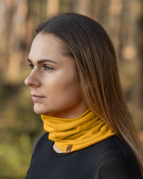 A woman with long, straight light brown hair is shown in a profile view, looking towards the left. She is wearing a spicy yellow menique neck gaiter and a black long-sleeved shirt, with a blurred background suggesting an outdoor, wooded environment.