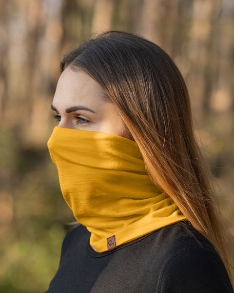 A woman with long, straight light brown hair is shown in a profile view, looking towards the left. She is wearing a spicy yellow menique neck gaiter and a black long-sleeved shirt, with a blurred background suggesting an outdoor, wooded environment.