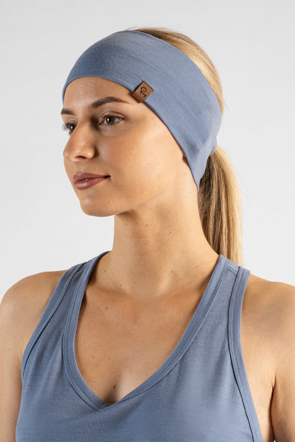 A close-up studio portrait of a blonde woman shown in a three-quarter profile looking off to the left against a plain light grey background. She is wearing a dusty blue sleeveless sport top and a matching wide headband featuring a small brown logo tag, with her hair pulled back into a ponytail.
