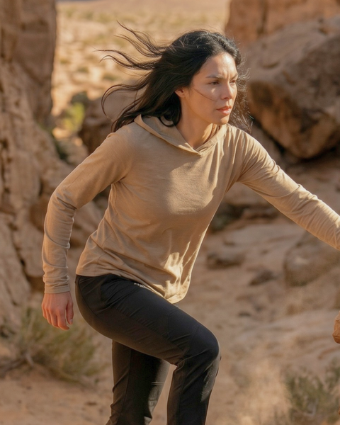 An outdoor shot of a woman with dark hair blowing in the wind, actively stepping up rocky terrain in a desert canyon setting. She is wearing a lightweight beige long-sleeved hoodie with the hood down and black pants.